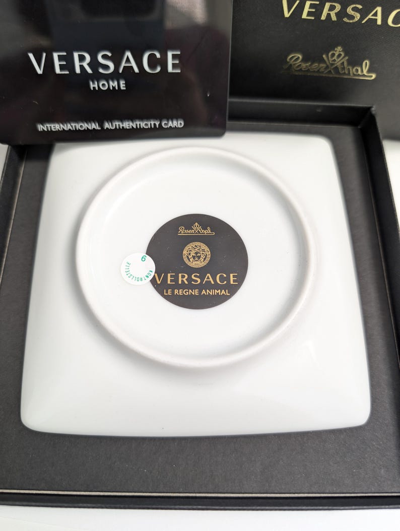 Puede incluir: Un plato cuadrado de porcelana blanca con una hendidura circular en el centro. El plato tiene el texto "VERSACE LE REGNE ANIMAL" y un emblema dorado. Se encuentra en una caja negra con una tarjeta de autenticidad Versace Home.