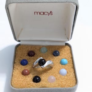 May include: A silver ring with a black stone, displayed in a Macy's box with various colorful gemstones. The ring has a unique, flowing design. The box is gray with a tan interior.