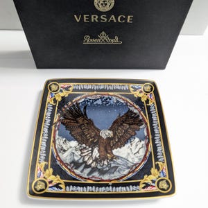 Puede incluir: Plato decorativo cuadrado con una ilustraci&oacute;n detallada de un &aacute;guila con las alas extendidas sobre un fondo de monta&ntilde;as nevadas. Borde negro con detalles dorados. Logotipos de Versace y Rosenthal.