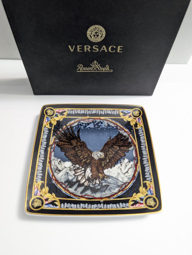 Puede incluir: Plato decorativo cuadrado con una ilustraci&oacute;n detallada de un &aacute;guila con las alas extendidas sobre un fondo de monta&ntilde;as nevadas. Borde negro con detalles dorados. Logotipos de Versace y Rosenthal.