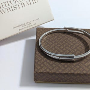 May include: A silver-toned metal bracelet with a unique overlapping design, resting on a brown patterned box. The box is labeled with the text "INTEREST WRISTBAND" and "AVON PRODUCTS INC."