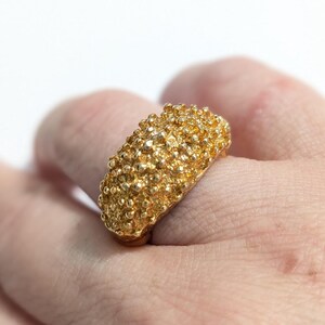 May include: A gold-colored ring with a textured surface covered in small, rounded bumps. The ring is held by a hand, showcasing its intricate design and metallic sheen. The ring's design is reminiscent of a cluster of tiny spheres.