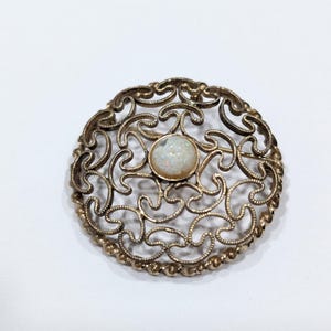 May include: A gold-toned filigree brooch with a white opal stone in the center.
