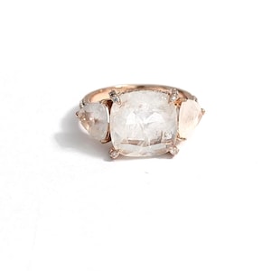 Könnte beinhalten: Ein Ring aus Roségold mit einem großen, mittig platzierten, kissenförmig geschliffenen, klaren Edelstein, flankiert von zwei kleineren, birnenförmigen klaren Edelsteinen. Das Band ist dünn und metallisch, mit kleinen Akzentsteinen an den Seiten.