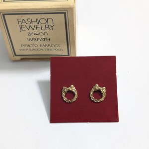 May include: Gold-toned wreath earrings with bow details on a red card. The earrings are displayed with their original box, which reads "Fashion Jewelry by Avon Wreath Pierced Earrings with Surgical Steel Posts."