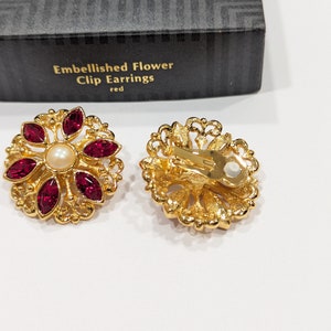 May include: A pair of gold tone clip-on earrings with a red flower design. Each earring features a faux pearl in the center and red rhinestones around the pearl. The earrings are in a box that says "Embellished Flower Clip Earrings red".