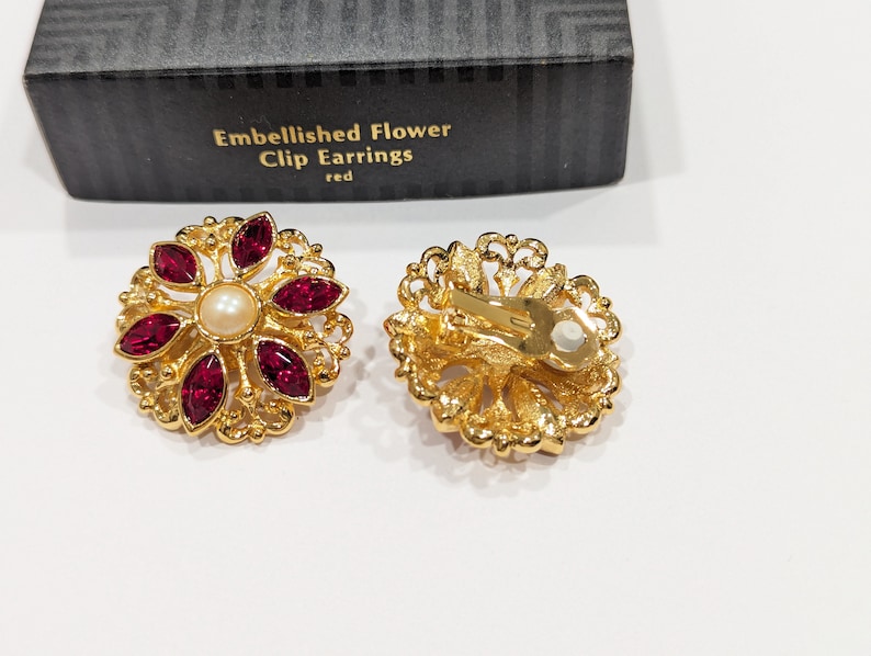 May include: A pair of gold tone clip-on earrings with a red flower design. Each earring features a faux pearl in the center and red rhinestones around the pearl. The earrings are in a box that says "Embellished Flower Clip Earrings red".