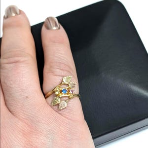 May include: A gold-colored ring featuring a leaf and branch design. The ring has a blue and pink gemstone. The ring is on a finger, with a black surface in the background.