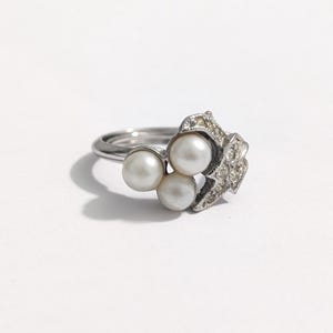 Op de afbeelding: Een zilverkleurige ring met drie witte parels als centraal element. De ring heeft een decoratief, sierlijk ontwerp met kleine, heldere stenen. De ring staat tegen een witte achtergrond.