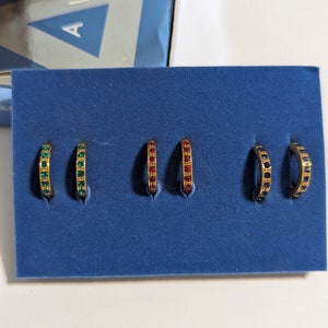 May include: A set of six gold hoop earrings with colorful gemstones. The earrings are displayed on a blue velvet background. The earrings are arranged in three pairs, with each pair featuring a different color of gemstone: green, red, and blue.
