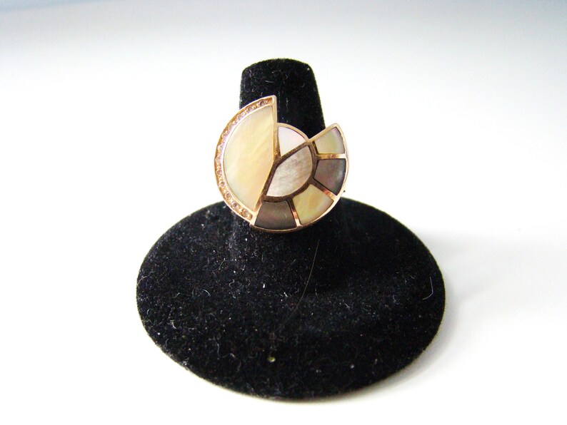 May include: A ring with a large, circular, multi-faceted gemstone set in gold. The gemstone is made of iridescent mother-of-pearl and has a geometric design.
