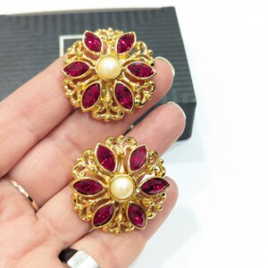 May include: A pair of gold tone earrings with a floral design. Each earring features a central pearl surrounded by eight red crystal stones.
