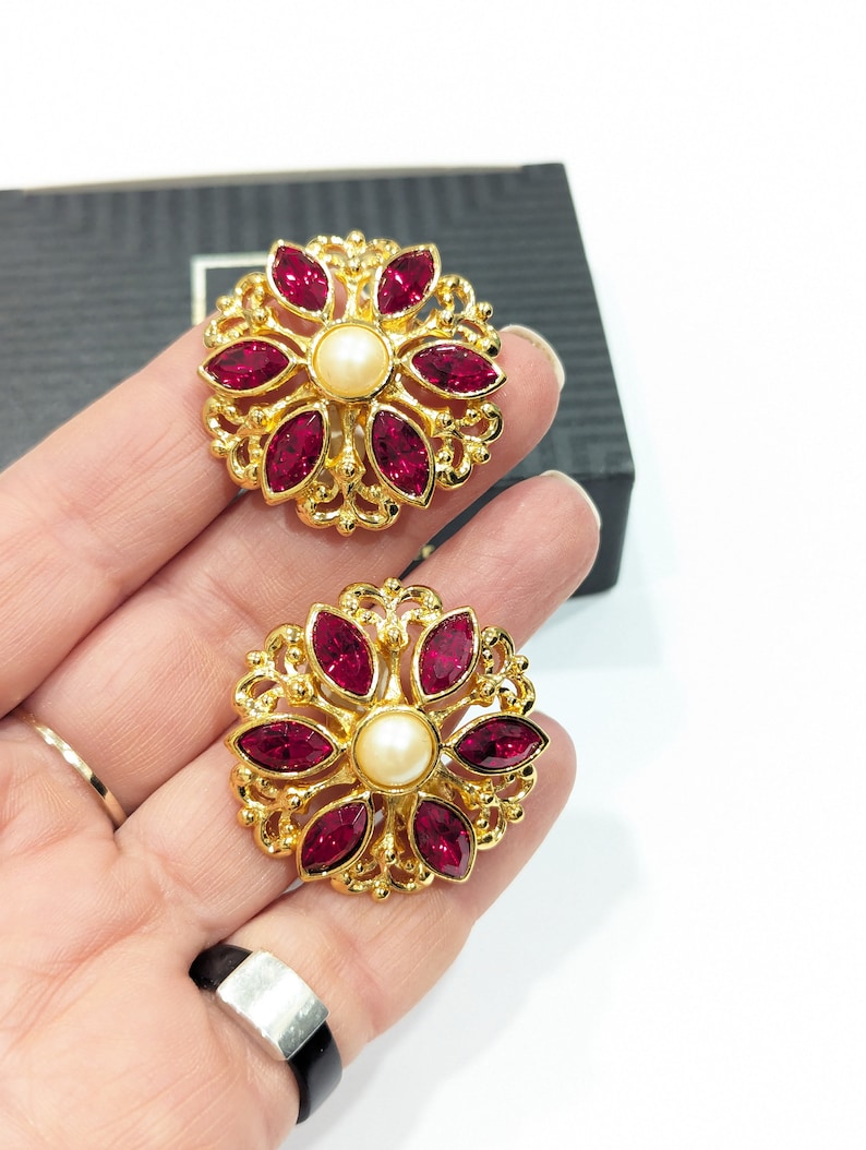 May include: A pair of gold tone earrings with a floral design. Each earring features a central pearl surrounded by eight red crystal stones.