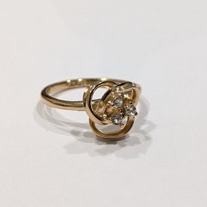 May include: A gold-coloured ring with a decorative floral design. The ring features a cluster of three small, clear gemstones at the centre of the design. The band is thin and simple, set against a white background.