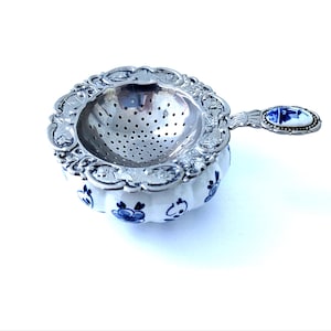 May include: A silver tea infuser with a white and blue floral patterned ceramic base. The infuser has a decorative handle with a blue and white design. The infuser is designed to hold loose leaf tea.