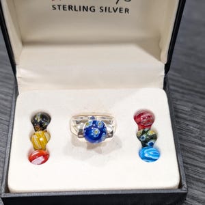 May include: A sterling silver ring with a large blue glass bead center, adorned with white floral designs. The ring is displayed in a black box with the text "STERLING SILVER".  Additional colorful glass beads are arranged on either side.