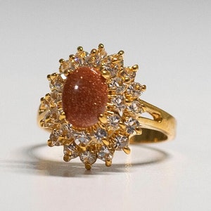 May include: A gold-toned ring featuring an oval, reddish-brown gemstone at the center, surrounded by a halo of clear, sparkling stones. The ring's band has a split design, adding to its elegant appearance. The setting is a sunburst style.