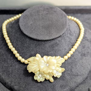 May include: A necklace featuring a strand of cream-coloured round beads. The centrepiece consists of a cluster of carved, off-white floral shapes made of a pearlescent material, accented with three small, clear, sparkling round rhinestones.
