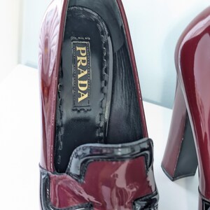 May include: Burgundy patent leather Prada loafers with a black trim and a chunky heel. The interior features the Prada logo in gold lettering. The shoes have a glossy finish and a classic design.