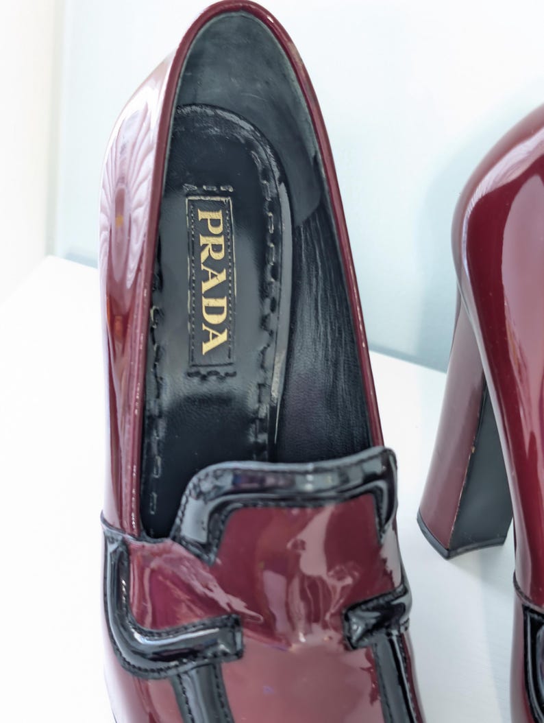 May include: Burgundy patent leather Prada loafers with a black trim and a chunky heel. The interior features the Prada logo in gold lettering. The shoes have a glossy finish and a classic design.