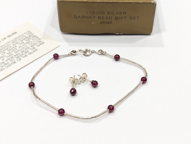 May include: A silver bracelet and earrings set with small red garnet beads. The jewelry is packaged in a box that reads "Liquid Silver Garnet Bead Gift Set, small".