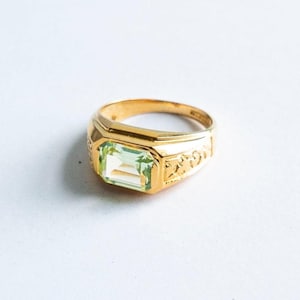 May include: A gold-toned ring with a rectangular, light green gemstone. The ring has a detailed design on the sides and a polished finish. The gemstone is set in a rectangular bezel.