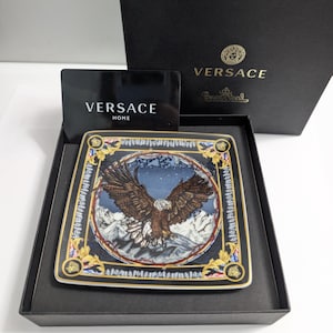 Puede incluir: Plato decorativo cuadrado con una ilustraci&oacute;n de un &aacute;guila sobre un fondo de monta&ntilde;as nevadas. Borde negro con detalles dorados. Presentado en una caja negra con el logo de Versace. Aproximadamente 20 cm x 20 cm.