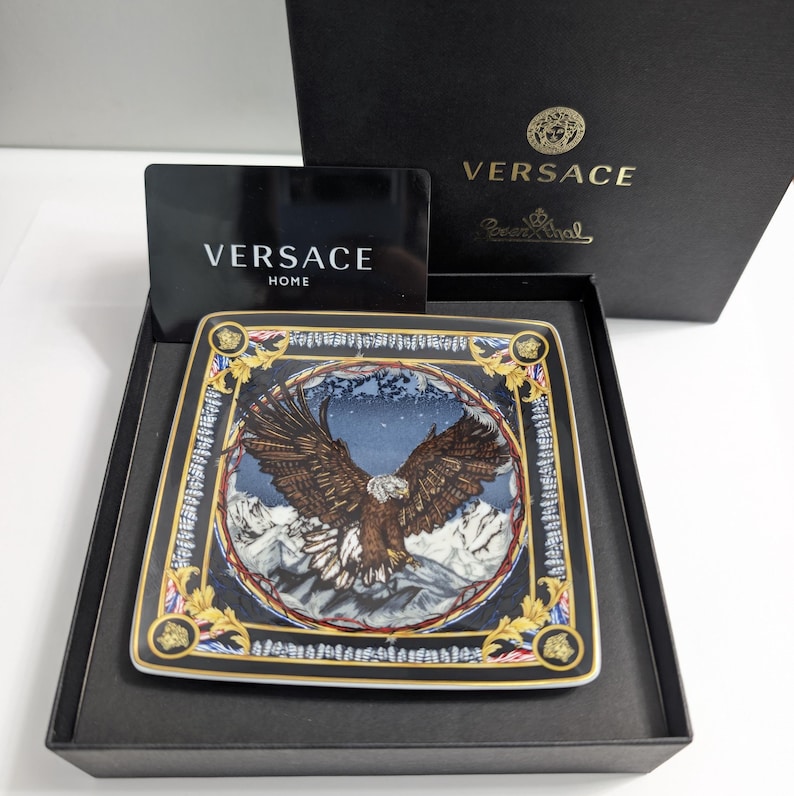 Puede incluir: Plato decorativo cuadrado con una ilustraci&oacute;n de un &aacute;guila sobre un fondo de monta&ntilde;as nevadas. Borde negro con detalles dorados. Presentado en una caja negra con el logo de Versace. Aproximadamente 20 cm x 20 cm.