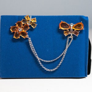 Può includere: Una collana a catena d'argento con due ciondoli: una farfalla con strass arancioni e trasparenti e un fiore con strass arancioni.
