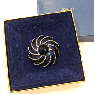 May include: A circular brooch featuring a central black cabochon surrounded by swirling, rope-textured silver-tone metal rays. The brooch is presented inside a square blue velvet-lined box with the gold text AVON printed on the lid.