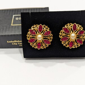 May include: A pair of gold clip-on earrings with a floral design. The earrings have a central pearl surrounded by red gemstones.