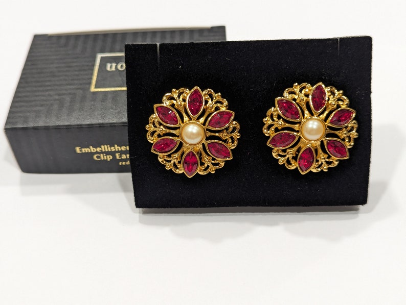 May include: A pair of gold clip-on earrings with a floral design. The earrings have a central pearl surrounded by red gemstones.