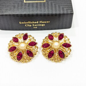May include: A pair of gold clip-on earrings with a red flower design and a pearl center. The earrings are labeled "Embellished Flower Clip Earrings red".