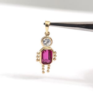 Peut inclure: Un pendentif en or en forme de personnage, doté d'une tête en pierre précieuse ronde et transparente et d'un torse en pierre précieuse rectangulaire rose foncé. Les bras et les jambes sont faits de petites perles d'or avec une bélière dorée.