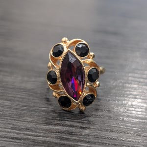 Puede incluir: Un anillo de oro con una gran piedra preciosa de color púrpura en forma de marquesa rodeada de piedras preciosas negras más pequeñas.