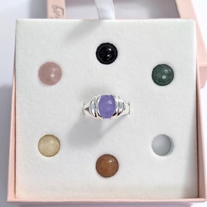 May include: A silver ring with a large, oval, lavender-colored stone is displayed in a pink box. The ring is surrounded by small, colorful, round stones in a white insert. The box is open, revealing the ring and stones.