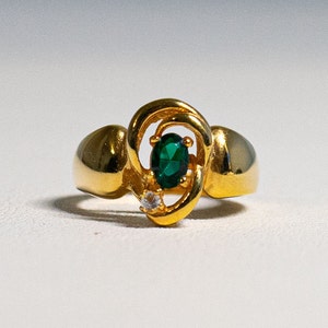 May include: A gold ring featuring a green oval-shaped gemstone and a smaller clear gemstone. The ring has a distinctive swirl design.