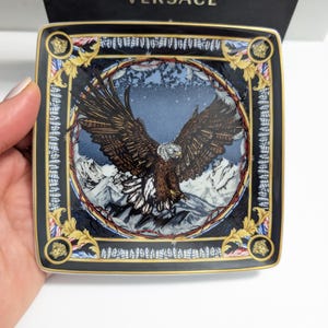 Puede incluir: Plato decorativo cuadrado con un dise&ntilde;o detallado de &aacute;guila sobre un fondo de monta&ntilde;as. El plato tiene un borde negro con detalles dorados y el logo de Versace. El plato est&aacute; sostenido por una mano.