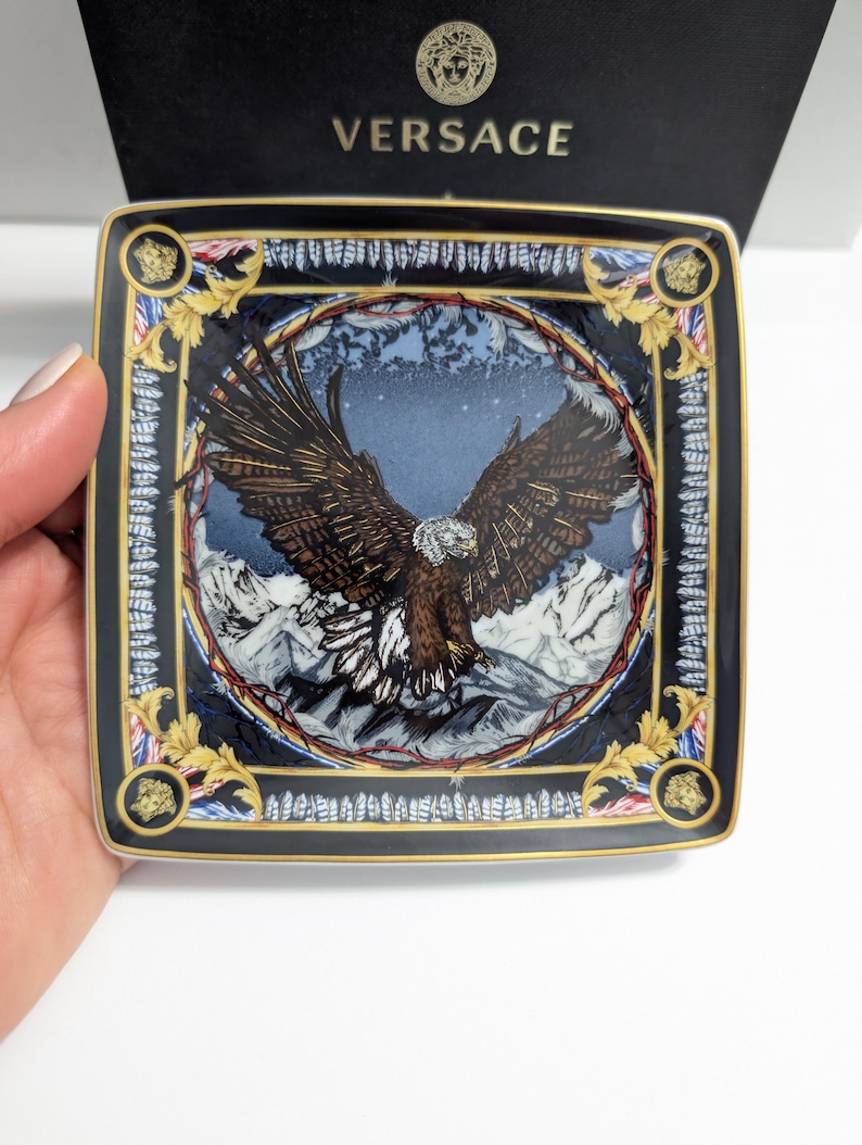 Puede incluir: Plato decorativo cuadrado con un dise&ntilde;o detallado de &aacute;guila sobre un fondo de monta&ntilde;as. El plato tiene un borde negro con detalles dorados y el logo de Versace. El plato est&aacute; sostenido por una mano.