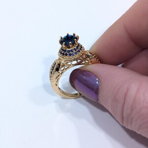 Peut inclure: Une bague en or avec une grande pierre pr&eacute;cieuse bleue entour&eacute;e de plus petites pierres bleues. La bague pr&eacute;sente un design d&eacute;licat et complexe.
