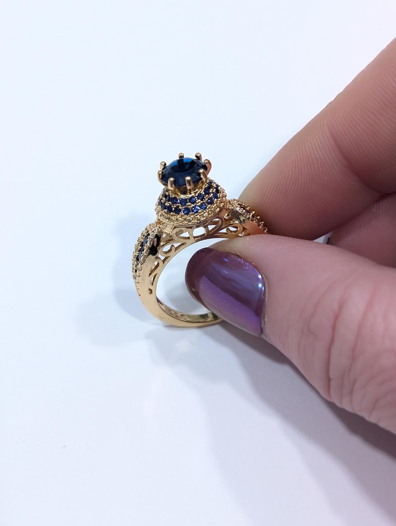 Peut inclure: Une bague en or avec une grande pierre pr&eacute;cieuse bleue entour&eacute;e de plus petites pierres bleues. La bague pr&eacute;sente un design d&eacute;licat et complexe.