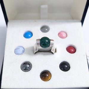 May include: A silver ring with a large, round, dark green stone at the center, surrounded by smaller, colorful gemstones. The ring is displayed in a white box with a dark blue border.