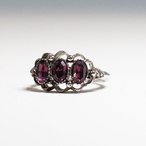 May include: A silver ring with three oval-shaped purple gemstones set in a delicate filigree design.