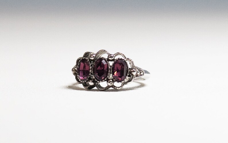 May include: A silver ring with three oval-shaped purple gemstones set in a delicate filigree design.