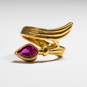 May include: A gold ring with a pink gemstone in the shape of a teardrop. The ring is designed with a curved band and a stylized wing shape.