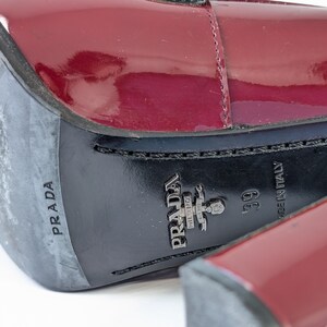 May include: Close-up of a pair of burgundy patent leather shoes. The soles are black with the brand name "PRADA" and "MADE IN ITALY" visible. The size is 39.