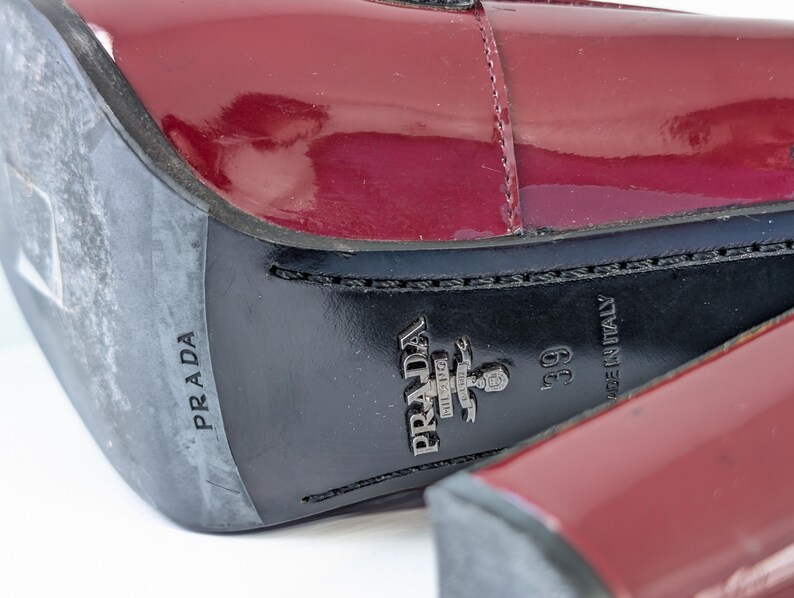May include: Close-up of a pair of burgundy patent leather shoes. The soles are black with the brand name "PRADA" and "MADE IN ITALY" visible. The size is 39.