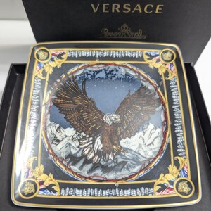 Puede incluir: Plato decorativo cuadrado con un dise&ntilde;o detallado de &aacute;guila, sobre un fondo de monta&ntilde;as. El plato presenta detalles dorados, un borde negro y los logotipos de Versace y Rosenthal. El plato se presenta en una caja negra.