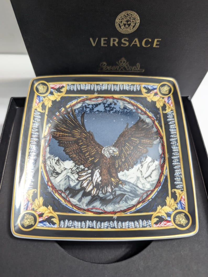 Puede incluir: Plato decorativo cuadrado con un dise&ntilde;o detallado de &aacute;guila, sobre un fondo de monta&ntilde;as. El plato presenta detalles dorados, un borde negro y los logotipos de Versace y Rosenthal. El plato se presenta en una caja negra.