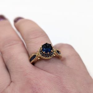 Peut inclure: Une bague en or avec une grande pierre ovale bleue entour&eacute;e de petites pierres bleues. La bague est port&eacute;e sur un doigt.